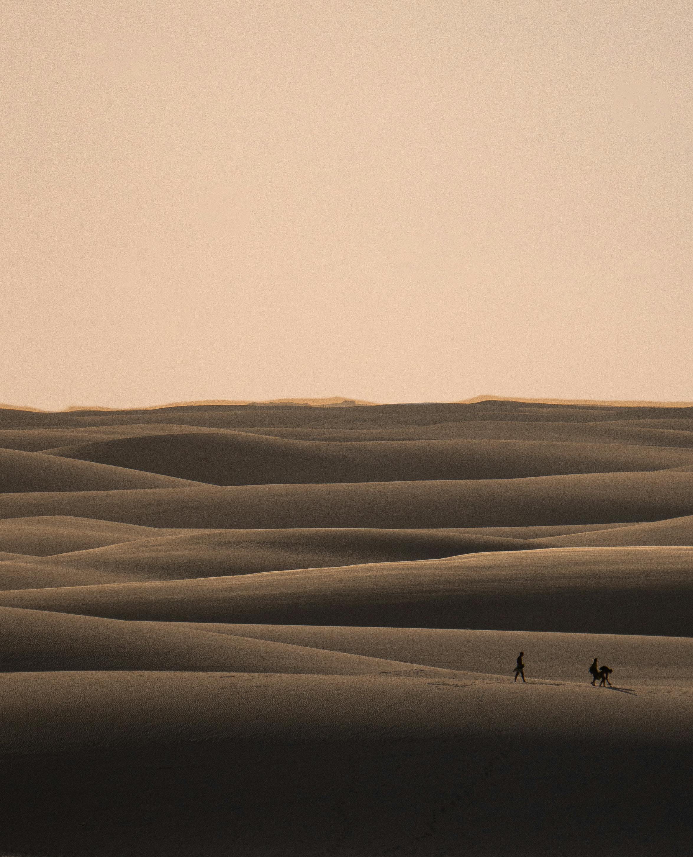 Two people walking on the vast serene sand dunes of Atins, Brazil under a clear sunset sky.