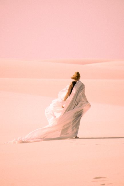 A woman wrapped in a flowing white shawl walks through desert sands in Atins, Brazil, under a pink sky.
