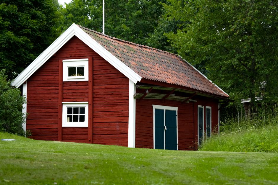 A quaint red wooden house nestled in a vibrant green countryside setting, surrounded by trees.