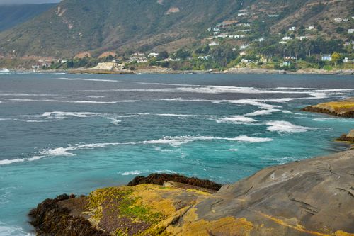 Free Tranquil coastal scene with moss-covered rocks, ocean waves, and hillside homes in the distance. Stock Photo