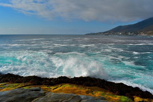Free Waves crash against colorful rocks on a scenic coastline with distant hills and cloudy sky. Stock Photo