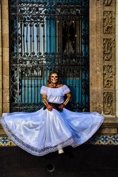 Woman celebrating Day of the Dead with traditional costume and makeup in front of ornate architecture.