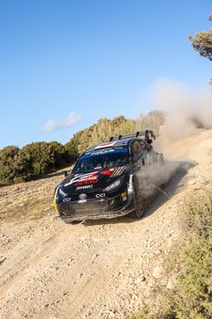 A Toyota Yaris GR racing car kicks up dust on a rugged dirt road in Sardinia, Italy.