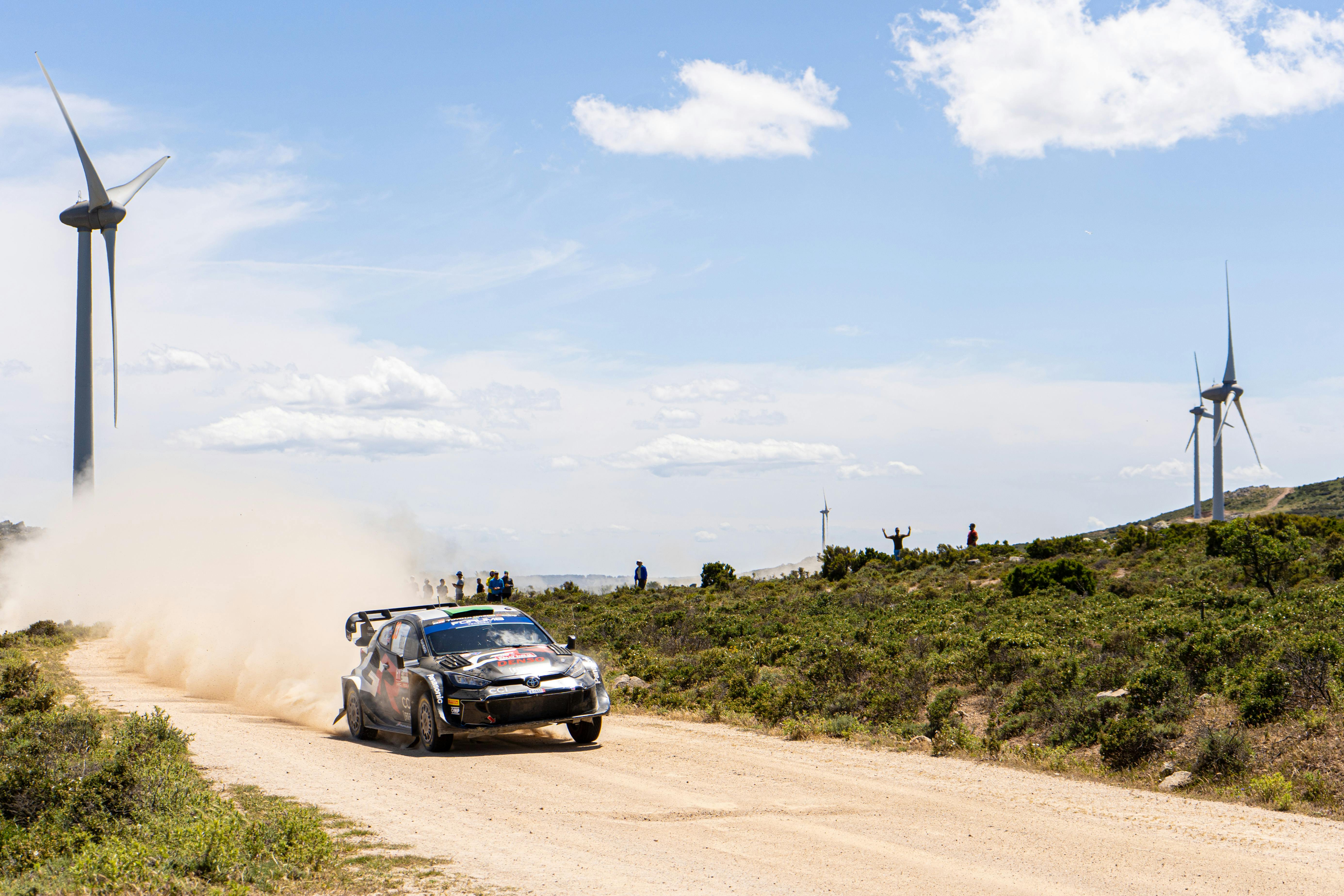 High-speed rally car racing past wind turbines on a dirt road in Sardinia.