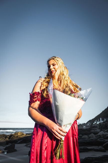 Elegant woman in a red dress with a bouquet standing by the beach under a clear blue sky.
