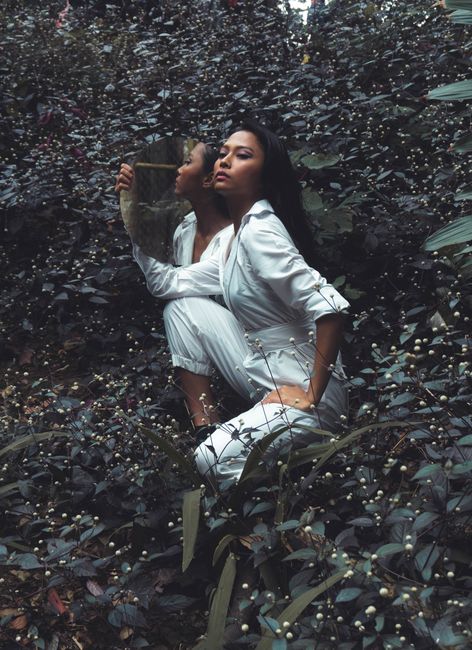 A stylish woman in white poses with a mirror amidst lush foliage, blending fashion with natural elements.