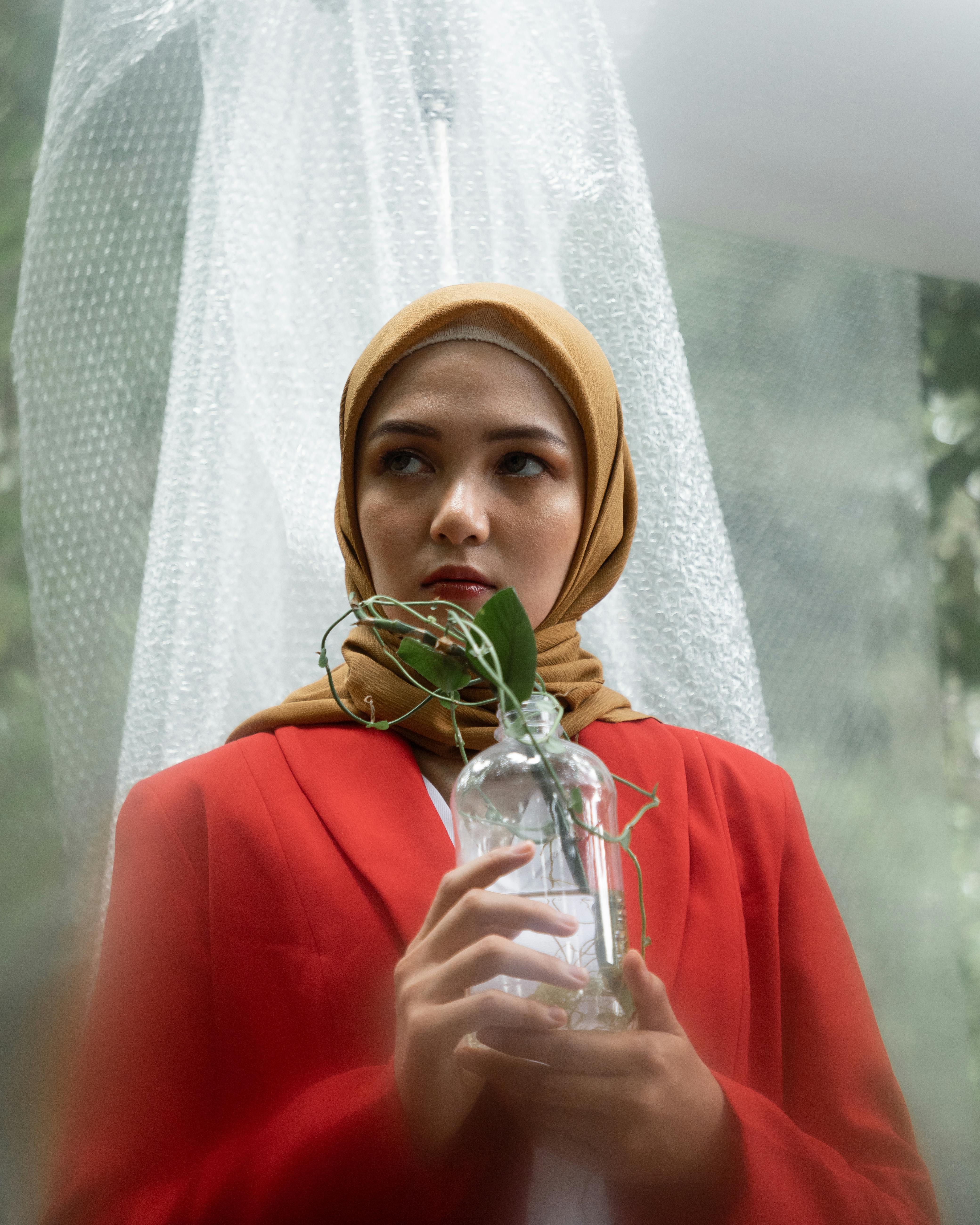 A woman in a red blazer and hijab holds a glass jar with a plant outdoors, symbolizing nature and elegance.