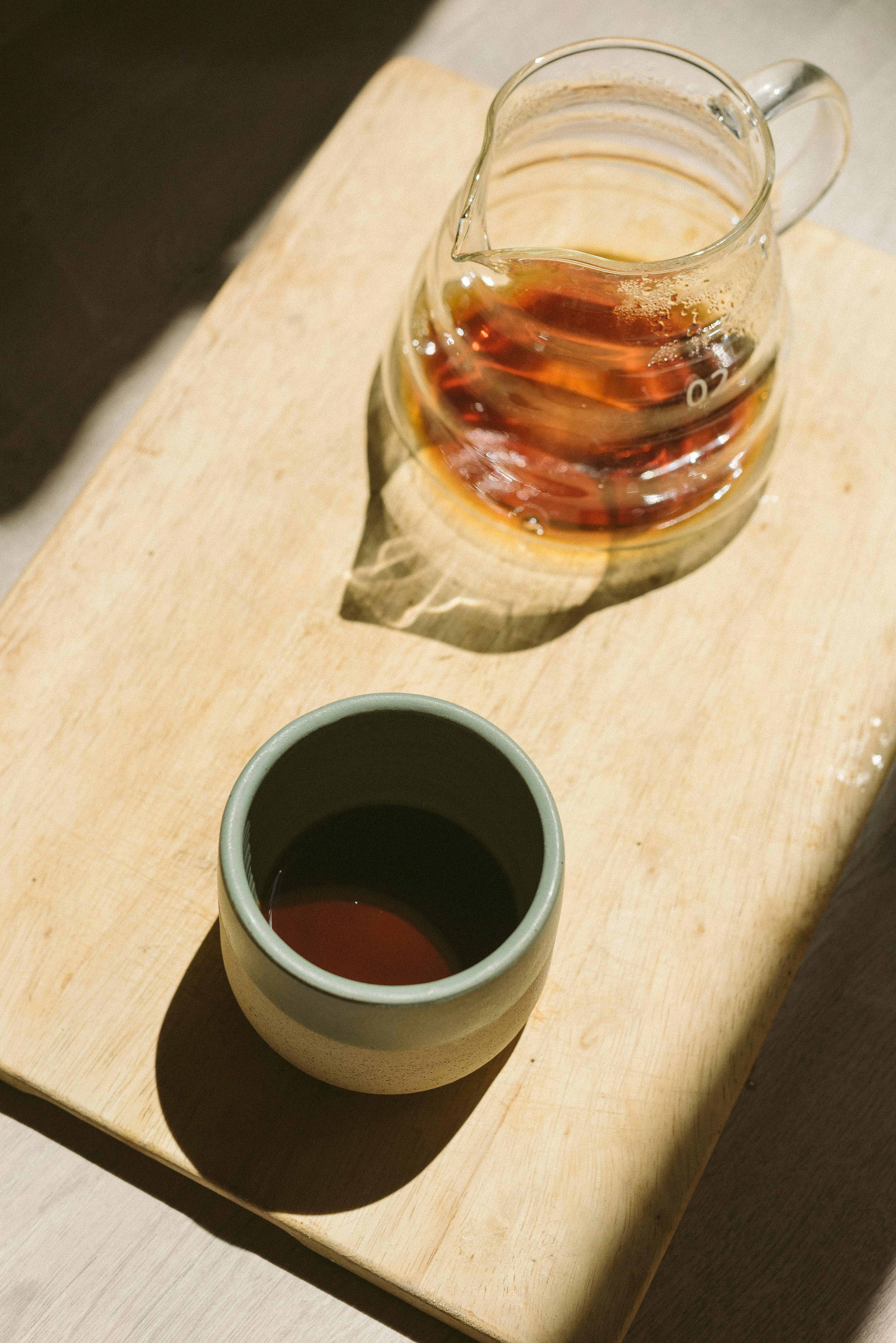 Glass pitcher and cup filled with tea on a wooden tray, bathed in sunlight.