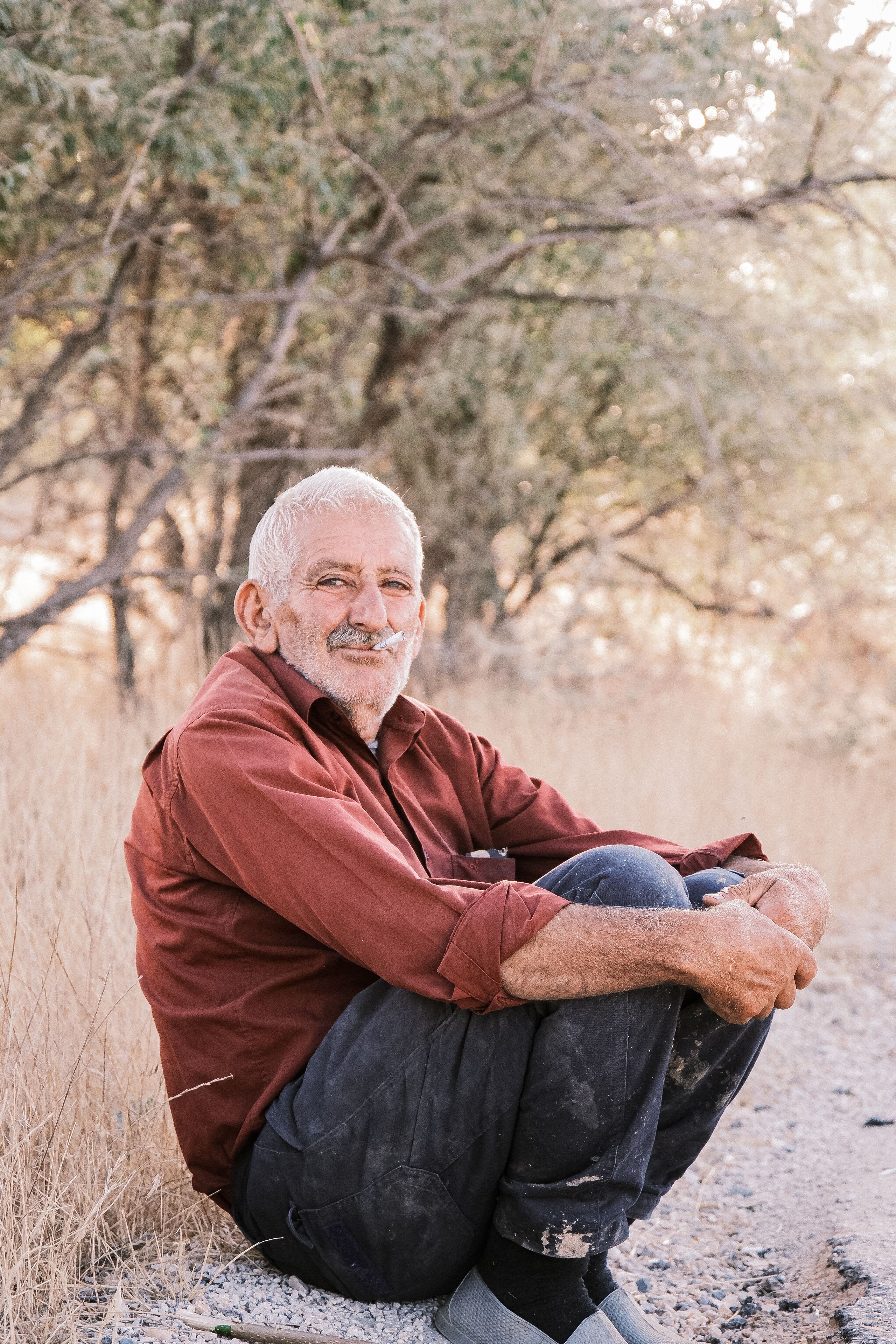 Elderly man sitting outdoors in a rural setting surrounded by nature, exuding calmness.
