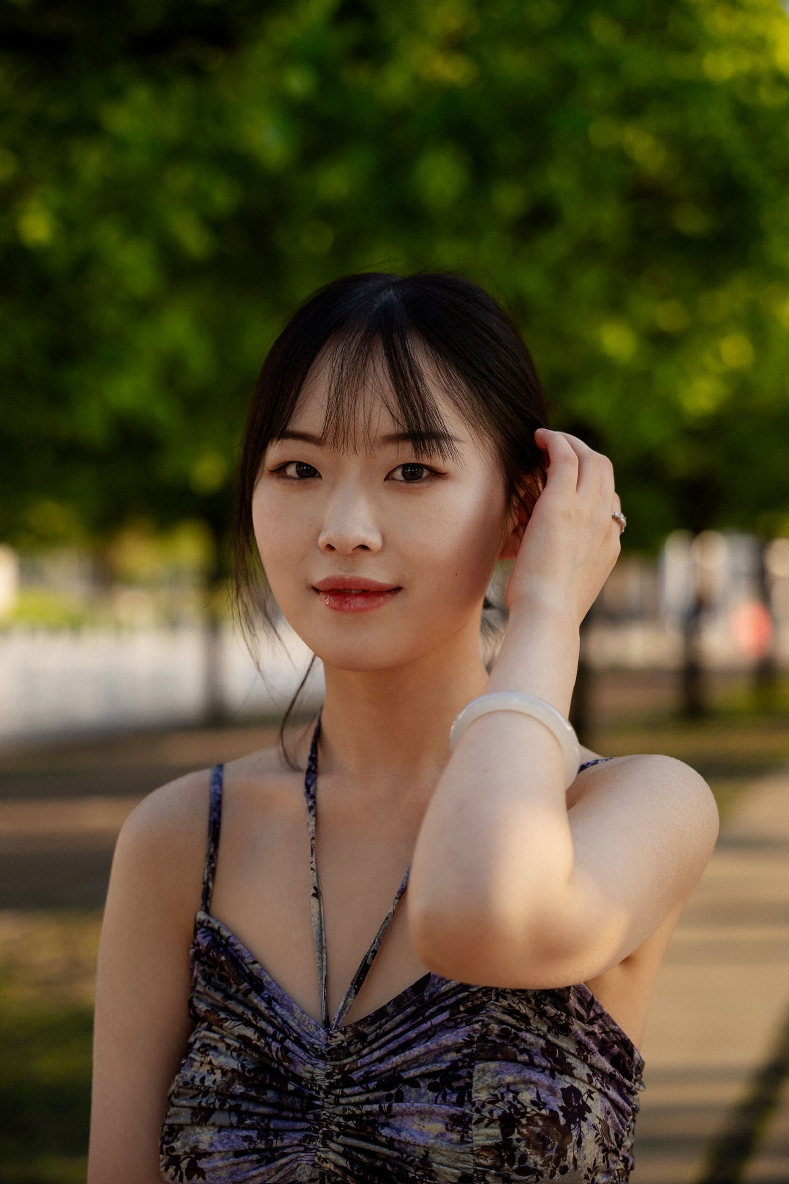 A young woman with bangs smiles gracefully in a sunny summer park.