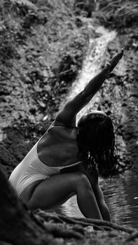 Woman practicing yoga in secluded natural setting near waterfall. Back view, black and white.