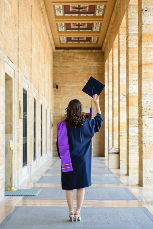 Free Young graduate celebrates in a grand architectural corridor, raising their cap in triumph. Stock Photo