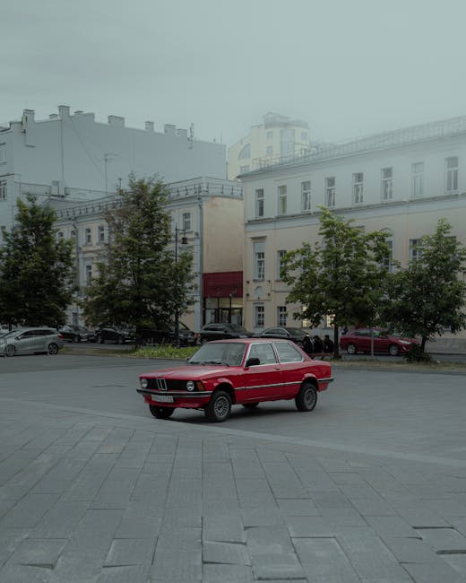 A vintage red car parked in a city square, surrounded by classic European architecture under an overcast sky.