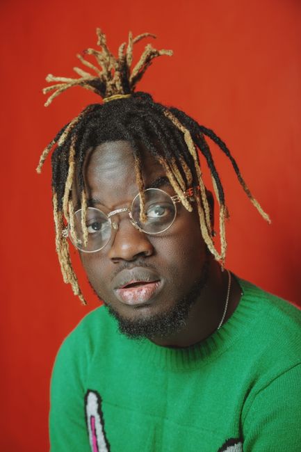 Portrait of a man with dreadlocks and glasses wearing a green sweater. Vibrant red background, showcasing contemporary style.