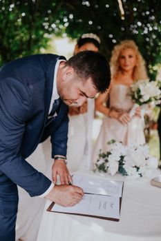 Groom signing marriage certificate with bride and witness during an outdoor wedding ceremony.
