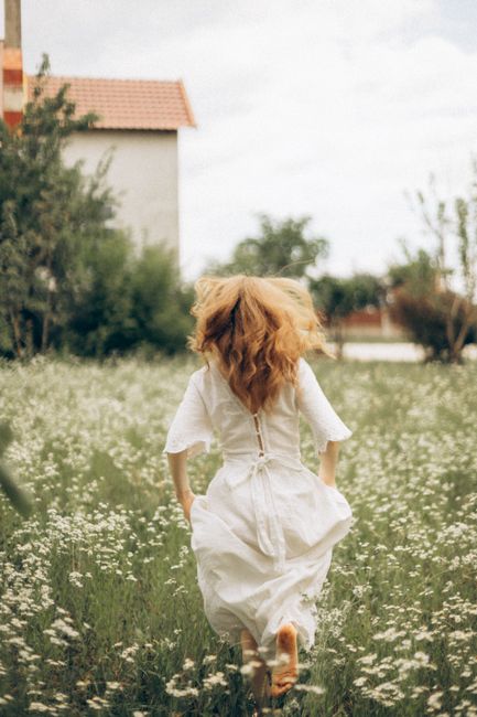 Woman in a white dress running barefoot through a flower-filled meadow on a bright day.