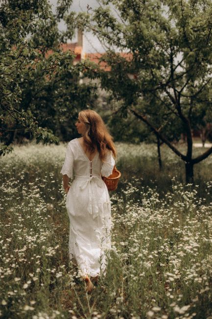 A woman in a white dress strolls through a flowering meadow carrying a wicker basket.
