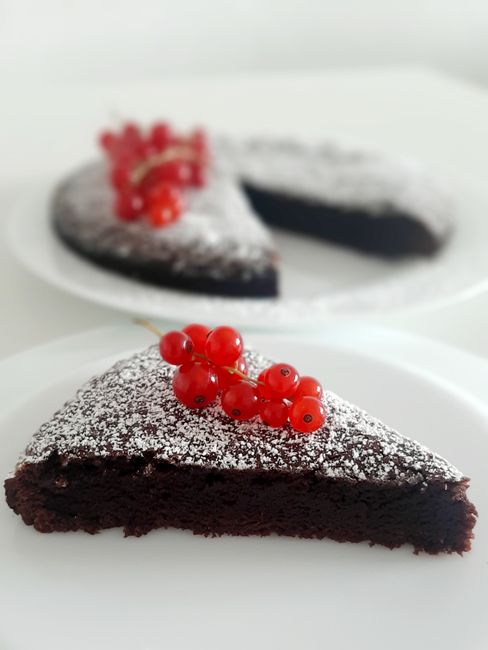 A sliced flourless chocolate cake on a white plate, showing a dense, fudgy interior and a crackled top dusted with powdered sugar.