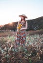 阳光明媚的田野里，一名身穿花卉连衣裙、戴着帽子的妇女在黄金时段捧着鲜花