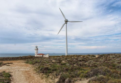 Free Panoramic view of Polente Lighthouse with a wind turbine by the sea under a vast blue sky. Stock Photo