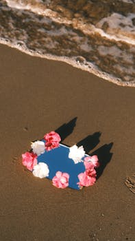 Mirror on beach sand with flowers, reflecting sky. Captures serene coastal beauty.