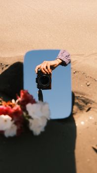 Artistic capture of a camera in a mirror reflection on a sandy beach with flowers.