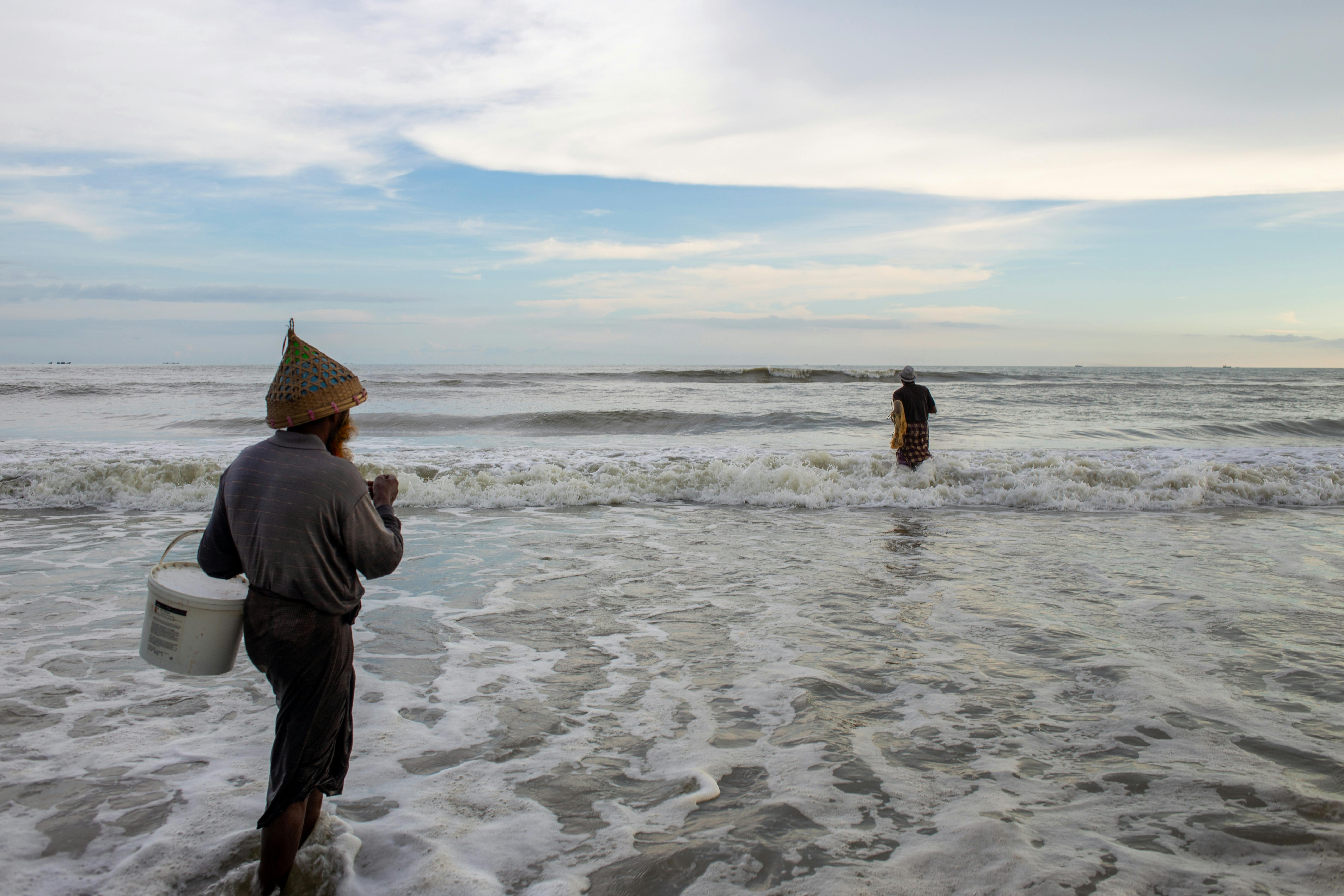 Two fishermen with buckets wade in the ocean waves at sunrise, wearing traditional hats.