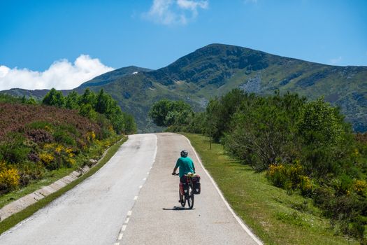 A cyclist rides along a scenic mountain road surrounded by lush greenery and blue skies.