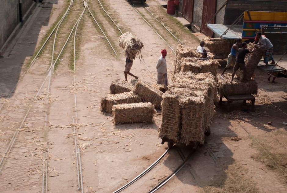 Men manually loading hay bales onto railway carts in a sunny outdoor rural area.