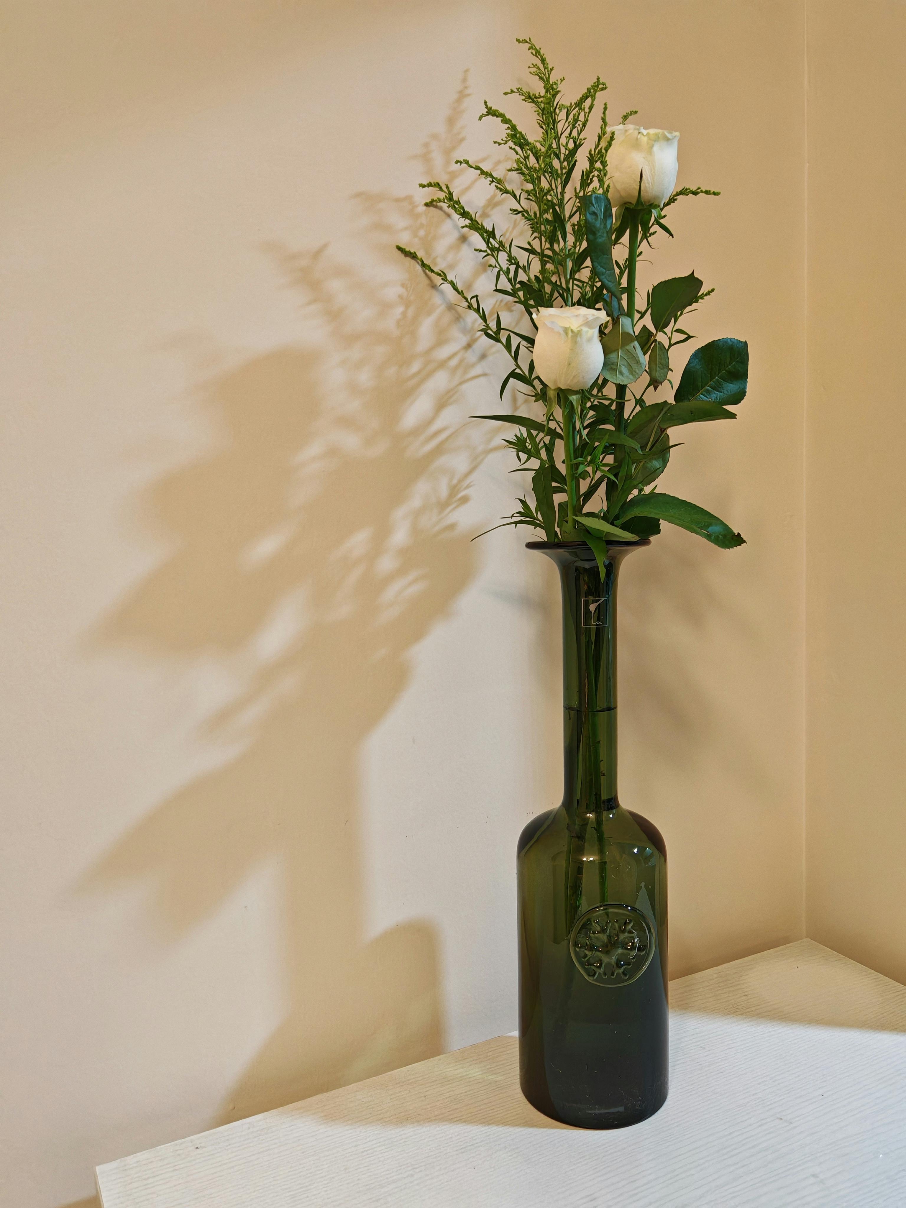 Free A minimalist floral arrangement with white roses in a green bottle vase casting shadows on a beige wall. Stock Photo