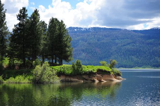A tranquil mountain lake scene featuring pine trees and hills under a cloudy sky.