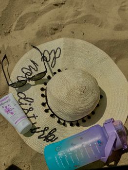 Flat lay of beach essentials: straw sun hat, sunglasses, sunscreen, and water bottle on sand.