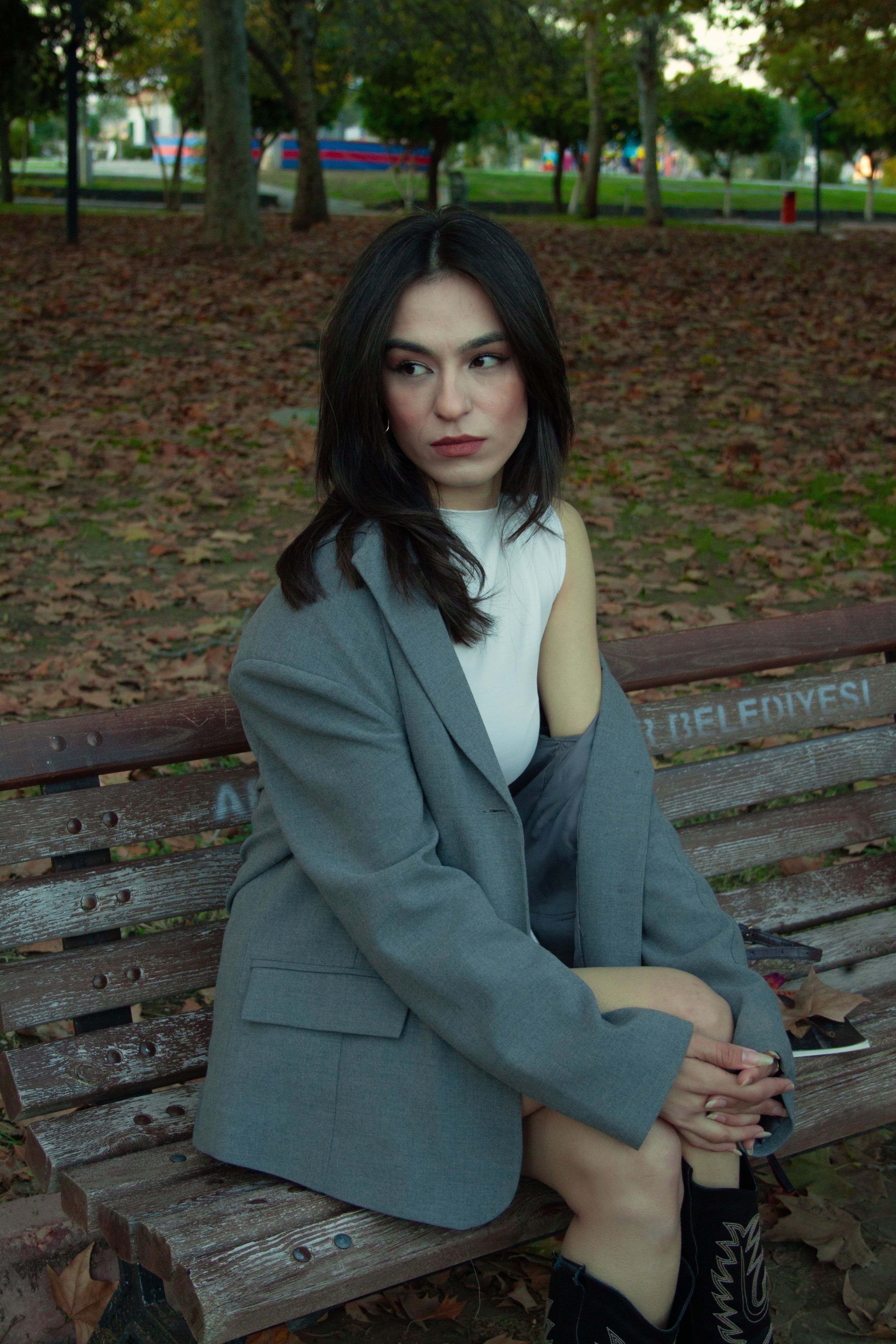 Fashionable woman in autumn attire sits on a park bench surrounded by fall leaves.