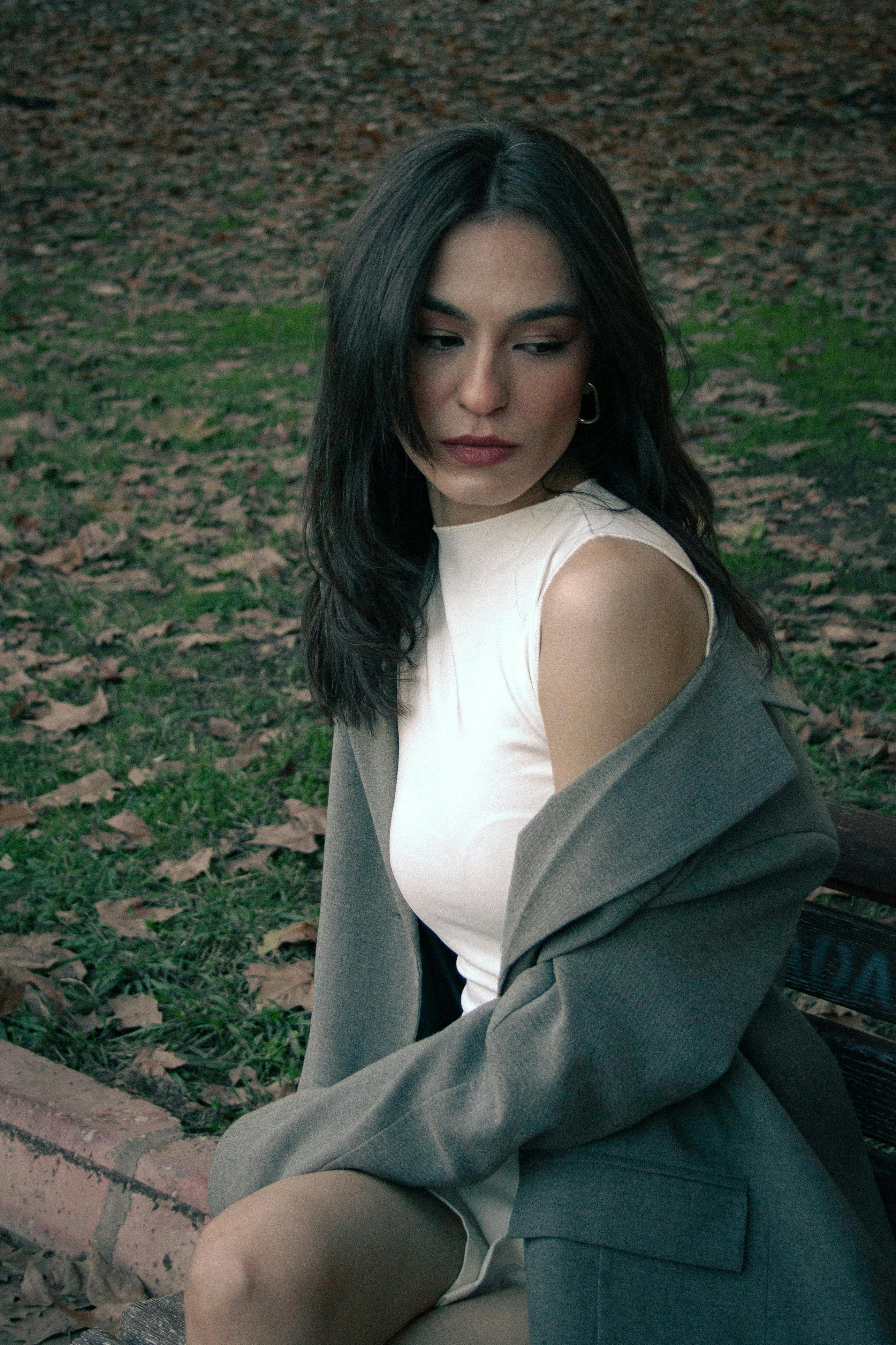 Elegant portrait of a woman in fall attire sitting on park bench, surrounded by fallen leaves.