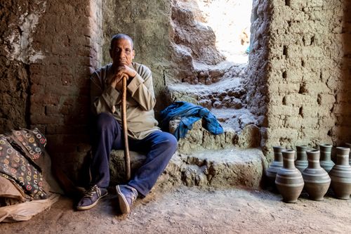 Free Elderly man holding a stick, sitting in a rustic, clay-walled interior surrounded by pottery and natural light. Stock Photo