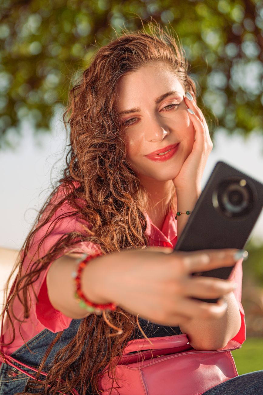 Smiling woman with long hair taking a selfie outdoors in Baghdad, Iraq, showcasing fashion and style.