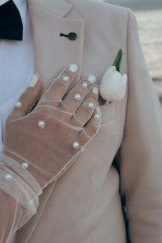 Close-up of bride's hand with pearl gloves on groom's suit adorned with white tulip.