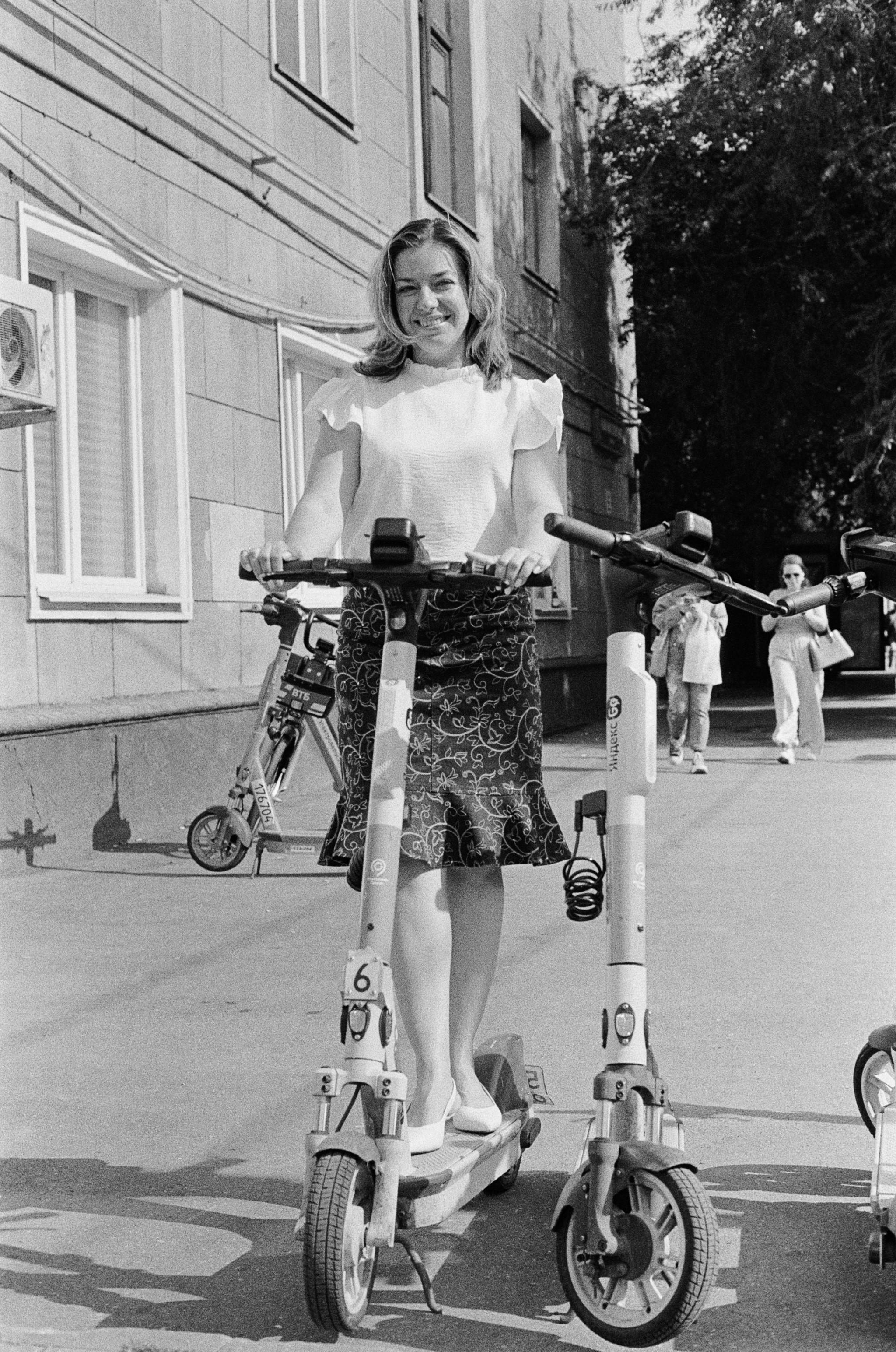 Free A woman in a skirt smiles while standing on an electric scooter on a sidewalk. Stock Photo