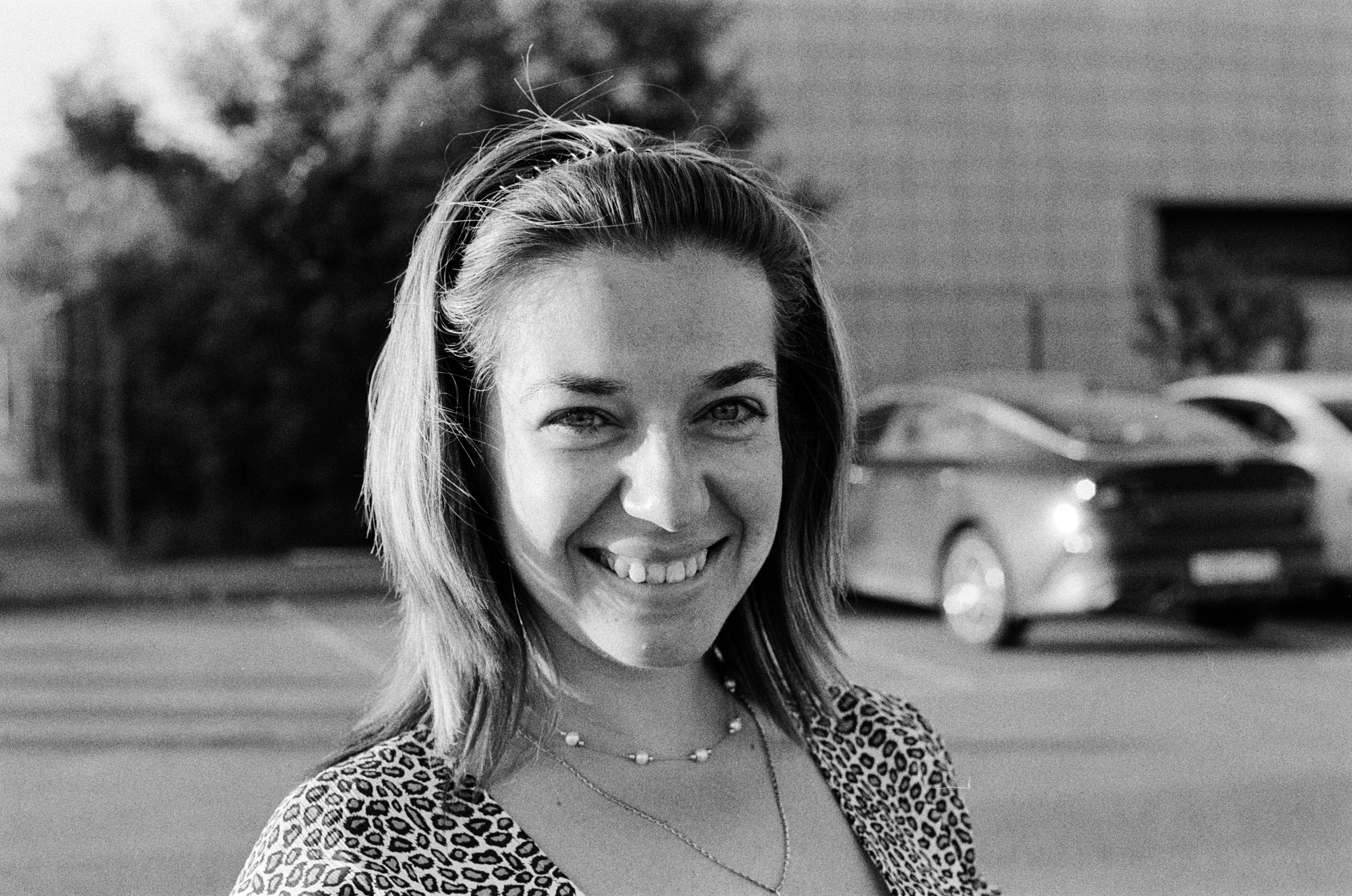 Black and white portrait of a smiling woman on a city street with cars in the background.