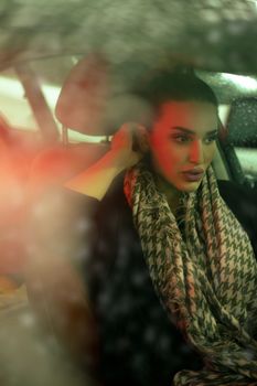 A sophisticated woman sits in a car, wrapped in a patterned gray scarf, exuding elegance and glamour.