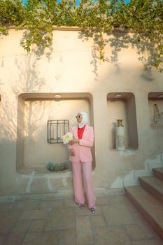 Elegant woman in pink suit with bouquet, posing outdoors against a wall in Baghdad.