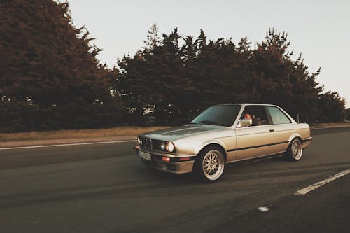 Vintage beige car with open window riding on empty asphalt road with trees behind in daytime