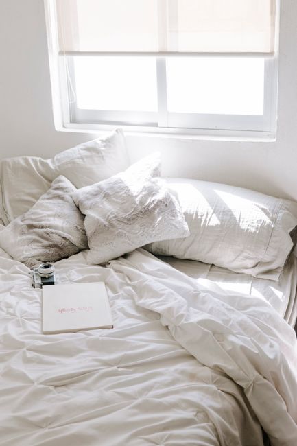 Bright and airy bedroom with rumpled bedding and pillows under a sunlit window.