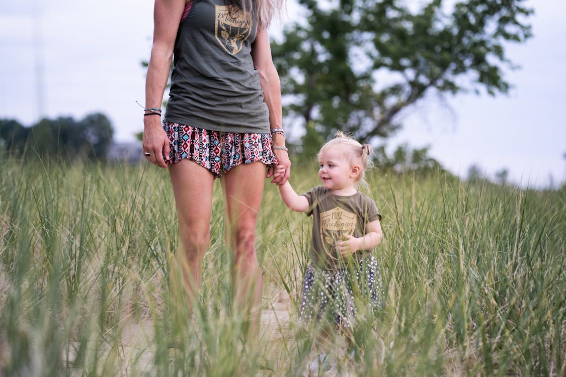 Free Woman Standing and Holding Hand of Baby Near Green Field Stock Photo