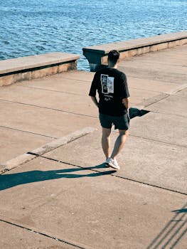 A man strolls along a sunny waterfront sidewalk in casual attire.
