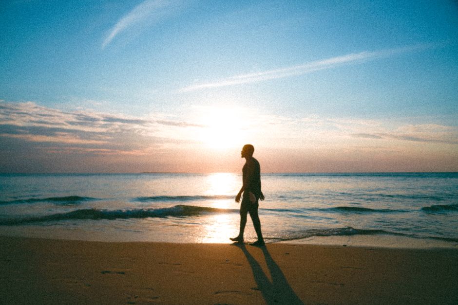 A man strolls along the beach at sunset with gentle waves and a serene sky.