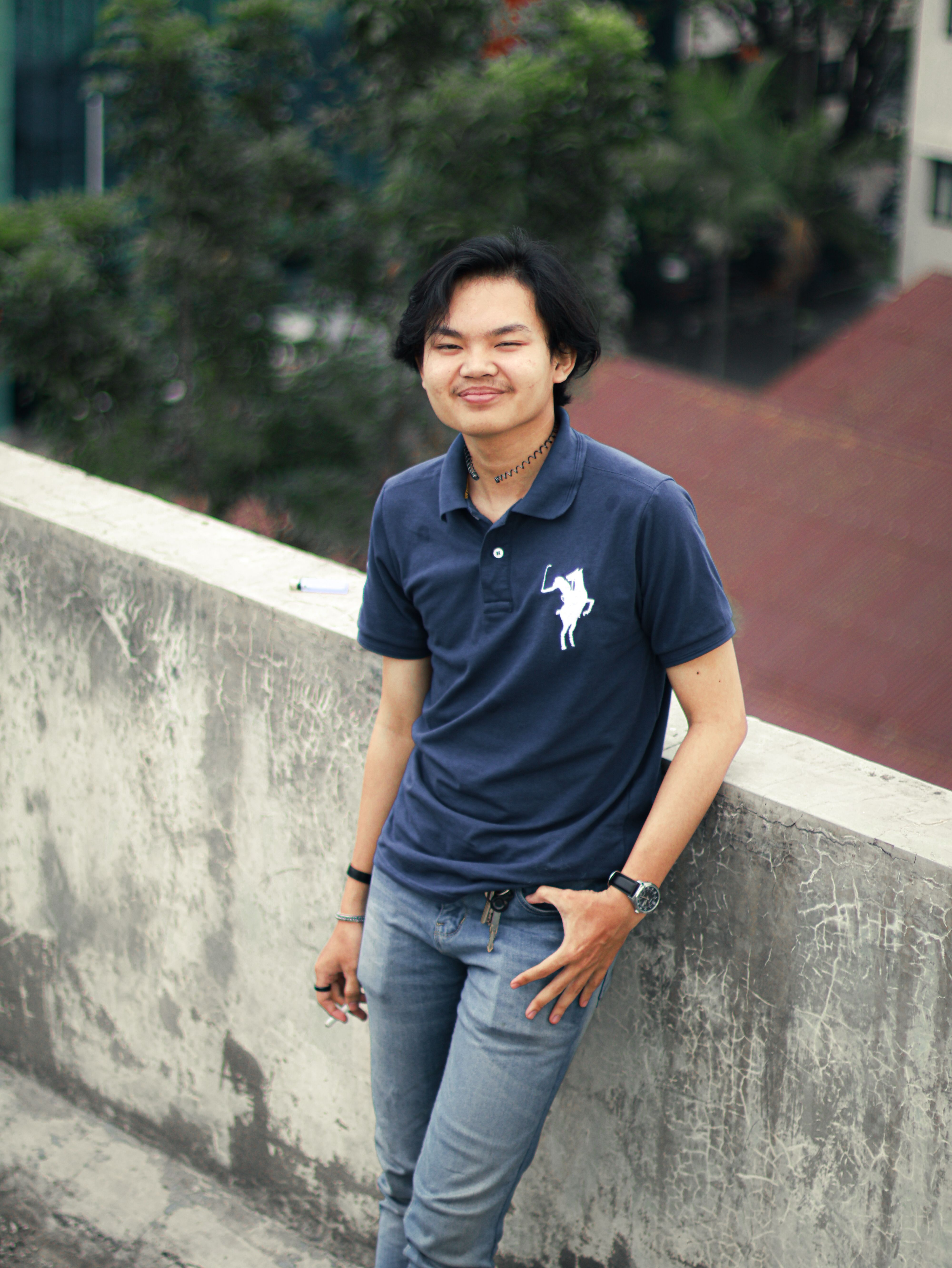 A young man in jeans and polo shirt is smiling while leaning on a concrete balcony outdoors.