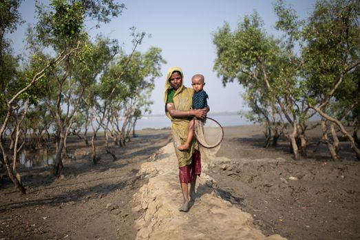 A South Asian woman walks holding her child through the muddy terrain of Sundarban, Bangladesh.