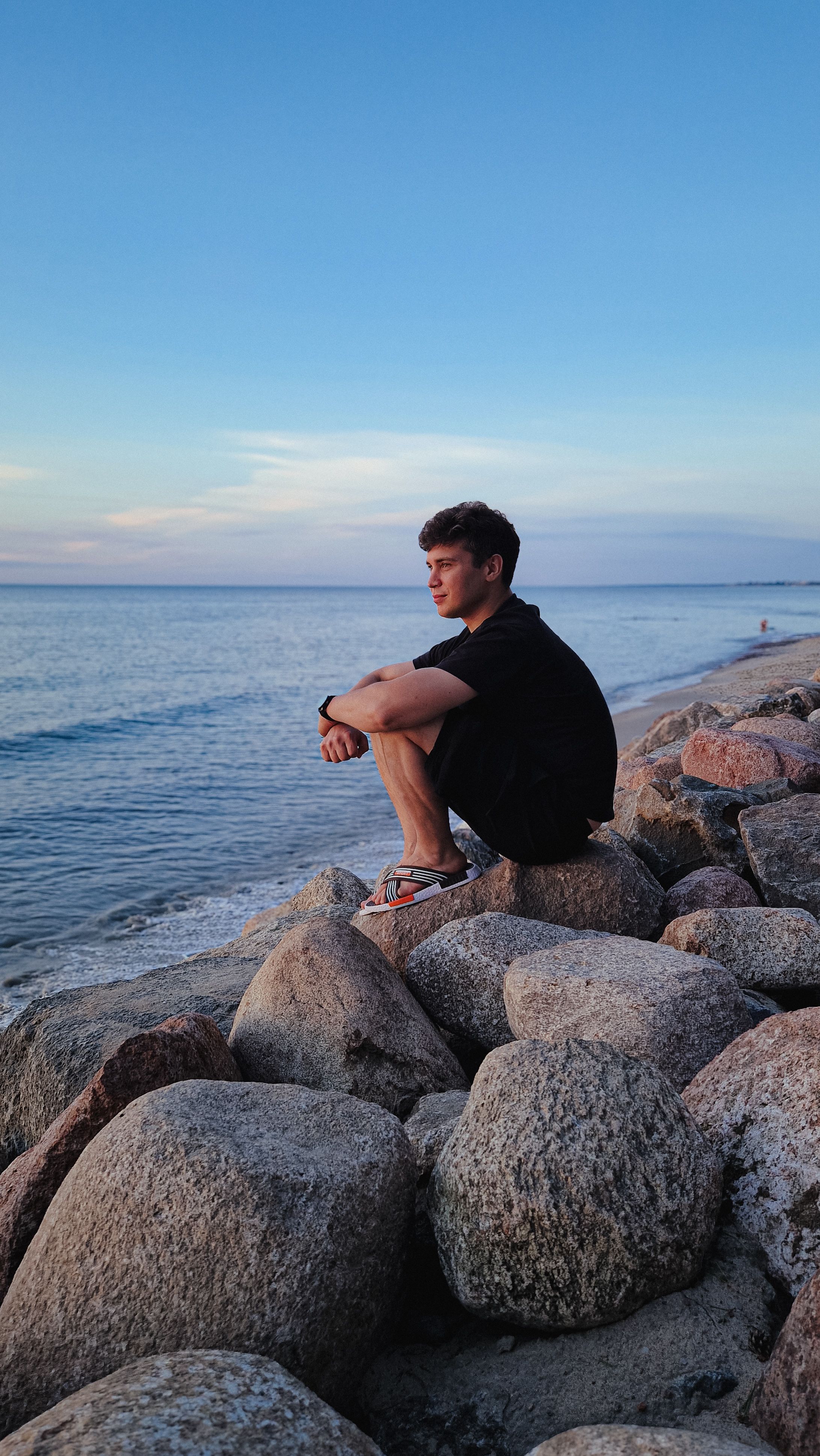 A man sits on rocks looking out at the calm sea during a tranquil summer evening.