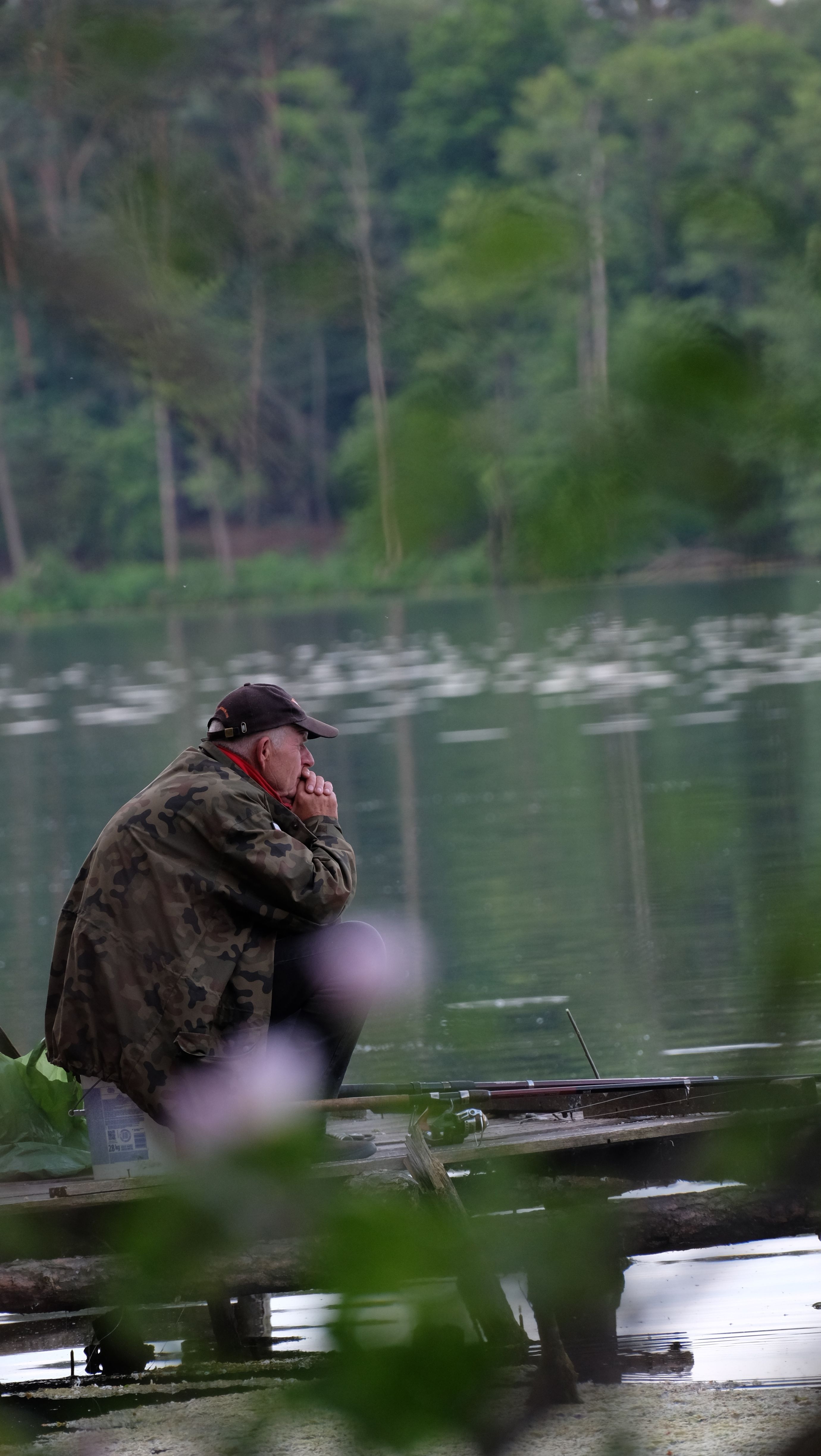 A man sits alone on a lakeside pier, fishing and pondering in nature.